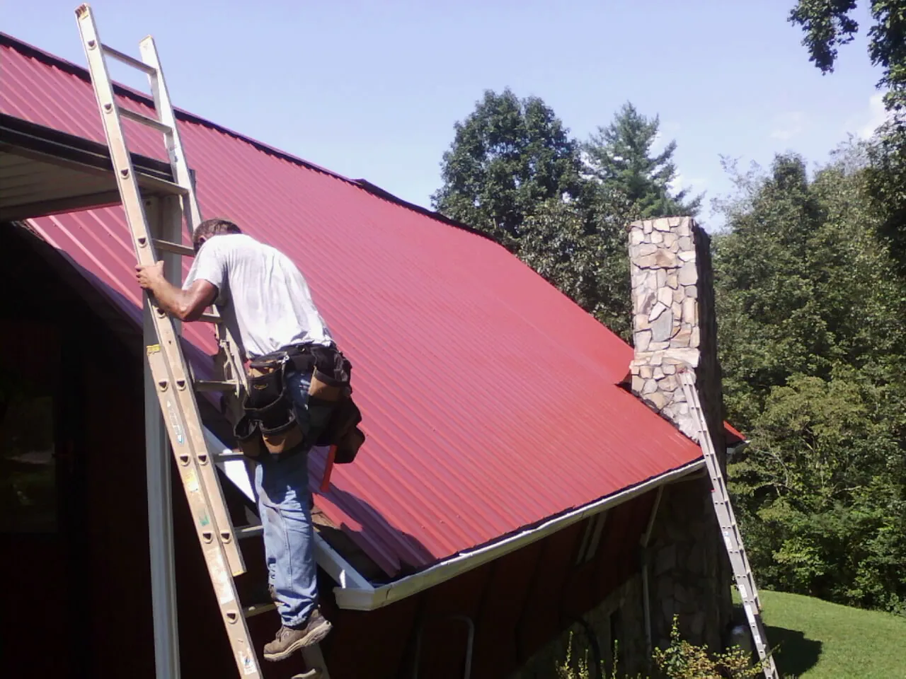 Roofer on a ladder beside a red metal roof and stone chimney — Soffit & Fascia Repair by Roofing Brothers in Horseheads